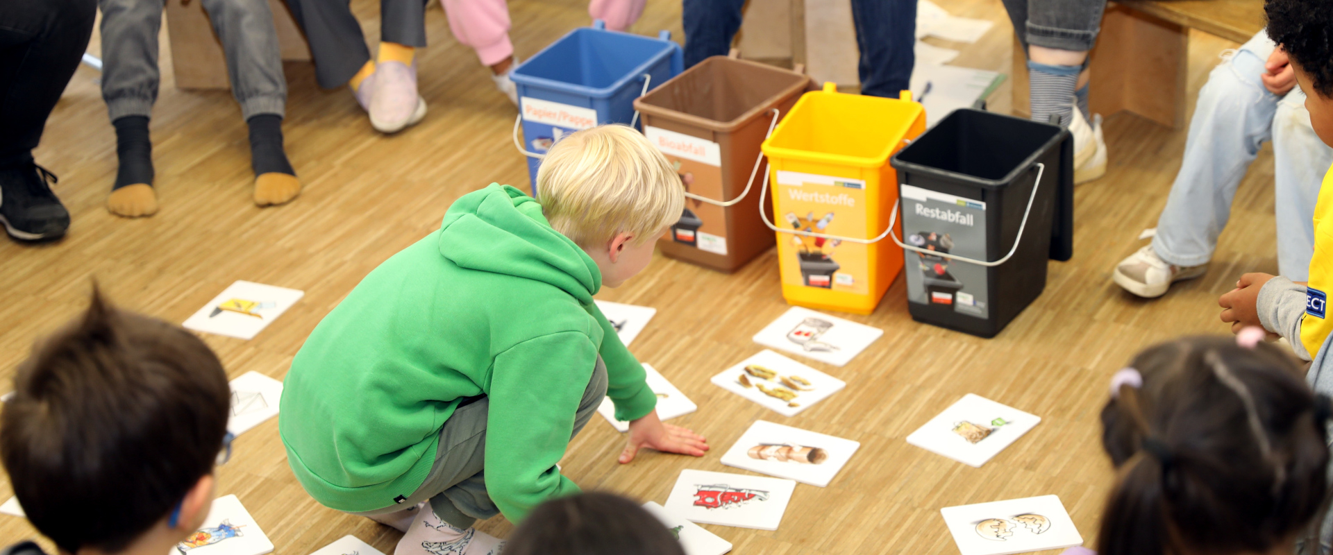 Kinder bilden einen Sitzkreis. Im Rahmen des Bildungsangebotes "Wertstoff:klasse" beschäftigt sich ein Kind in der Mitte des Stuhlkreises mit Lehrmaterialien.