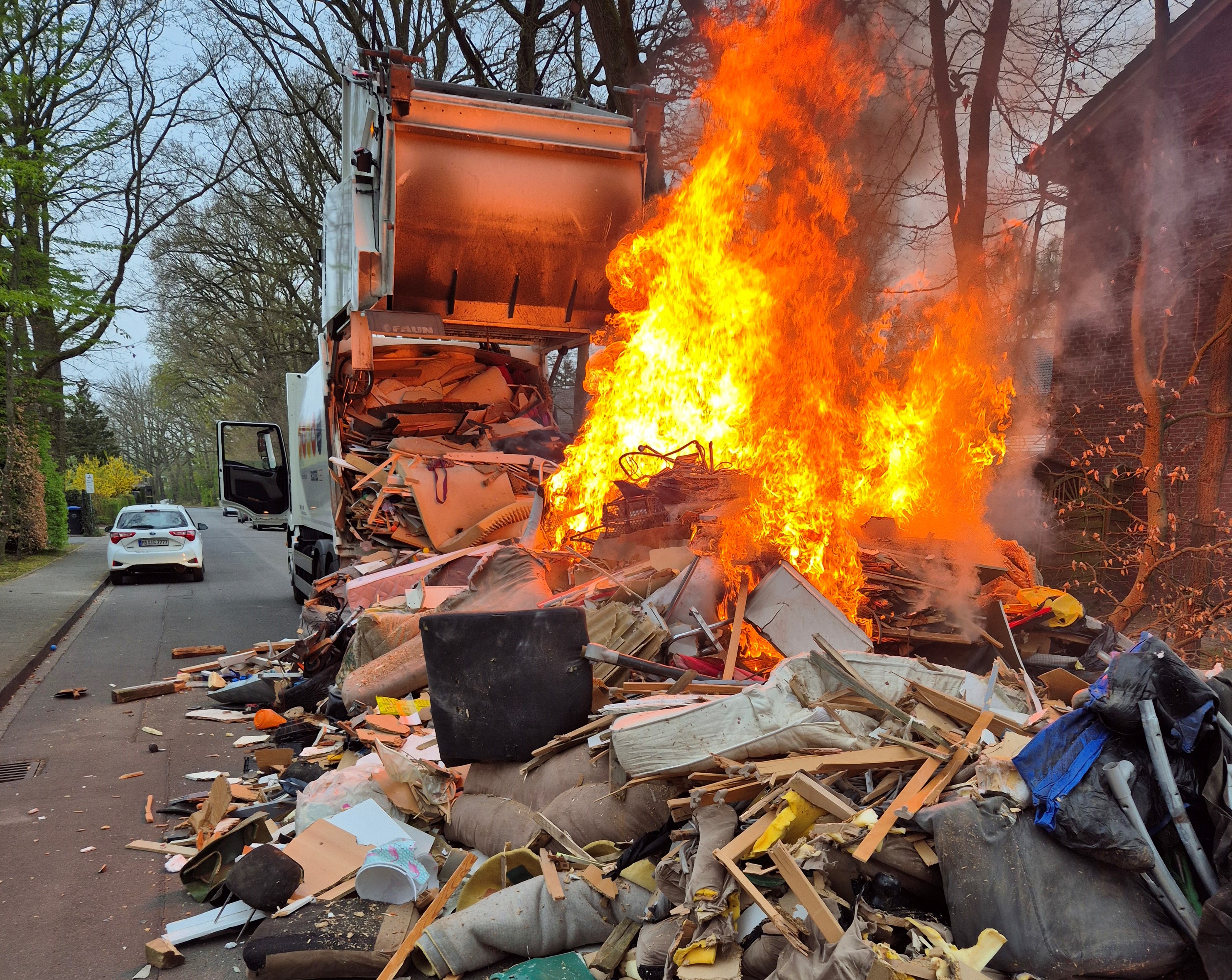Ein in Flammen stehender Sperrmüllhaufen, ausgeschüttet auf der Straße. Dahinter das Sperrgutfahrzeug mit geöffneter Heckklappe.